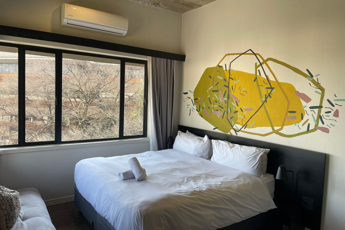 A modern bedroom featuring an exposed concrete ceiling, large window for natural light, wall-mounted air conditioning, and a custom built-in headboard.