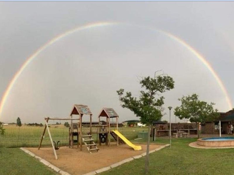 Expansive farm garden featuring a playground, swimming pool, and outdoor entertainment area under a double rainbow.