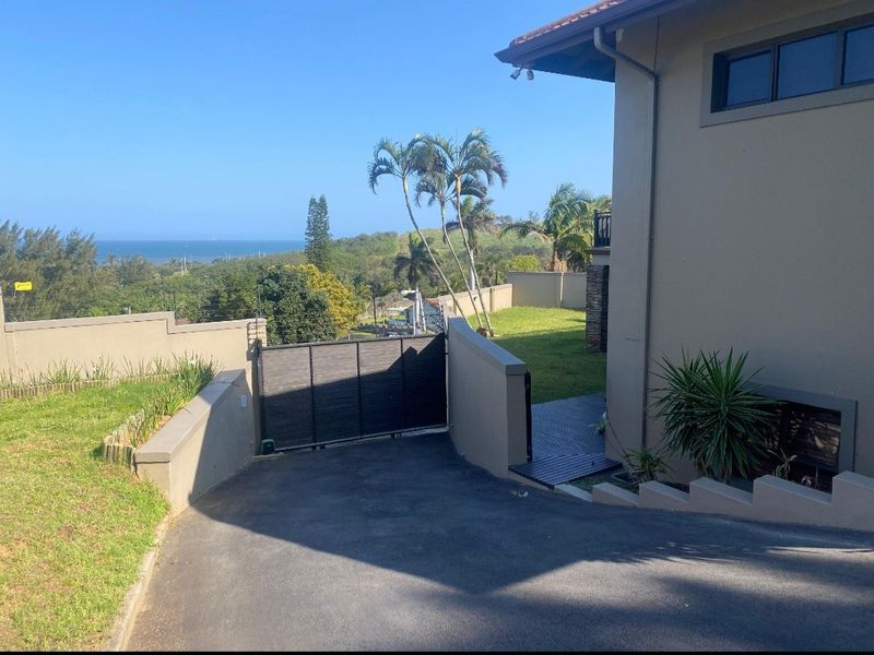Front exterior view featuring an asphalt driveway, secure metal gate, boundary walls, and a landscaped garden with scenic coastal views.