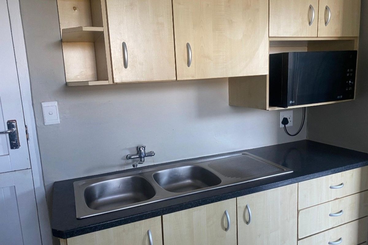 A functional kitchen area featuring light wood-tone cabinetry, a double stainless steel sink, and a dark laminate countertop with integrated microwave storage.