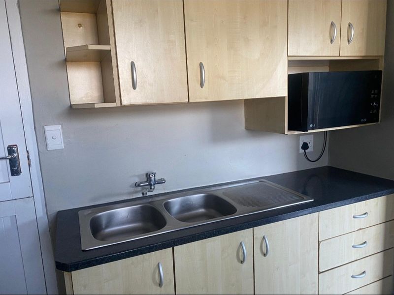 A functional kitchen area featuring light wood-tone cabinetry, a double stainless steel sink, and a dark laminate countertop with integrated microwave storage.
