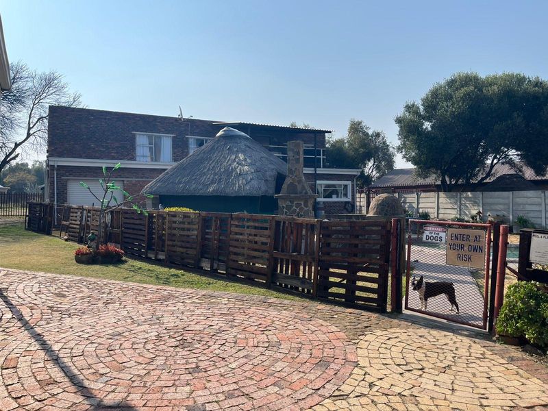 Rear exterior view featuring a thatched lapa, stone fireplace, swimming pool area, and brick paved driveway with perimeter fencing.