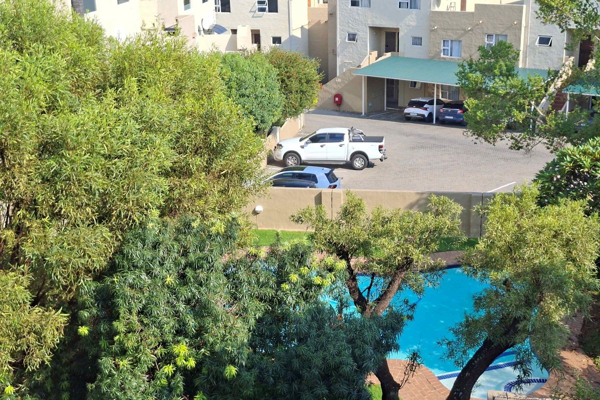 An elevated view of the apartment complex grounds, featuring a swimming pool, paved parking area, and established trees.
