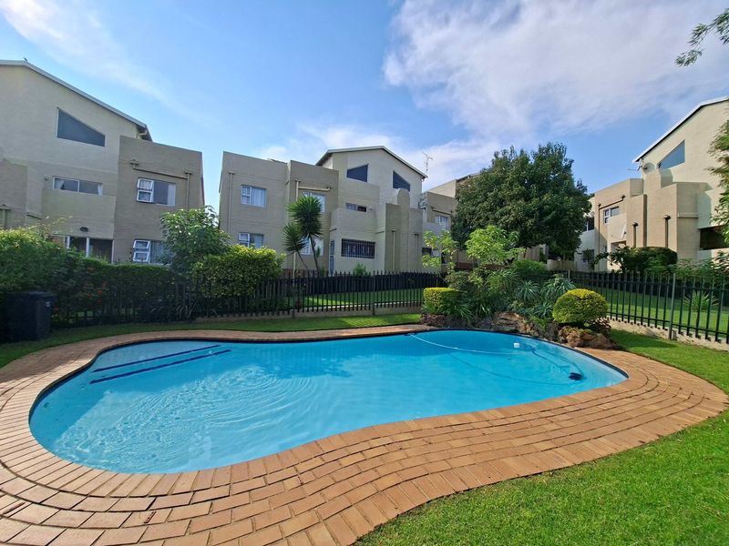 A well-maintained communal swimming pool area featuring brick coping, landscaped garden surrounds, and secure metal perimeter fencing within an apartment complex.