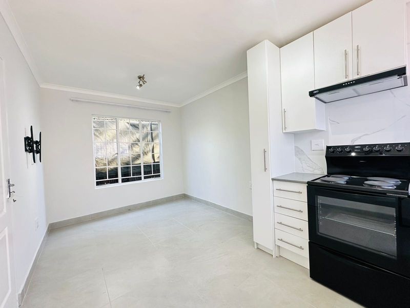 A bright, open-plan living and kitchen area featuring modern white cabinetry, a built-in stove with extractor, and neutral tiled flooring throughout.