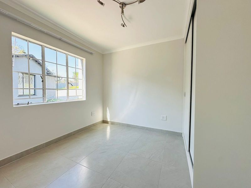 A bright, empty bedroom featuring neutral tiled flooring, built-in cupboards, and a large window with security bars.