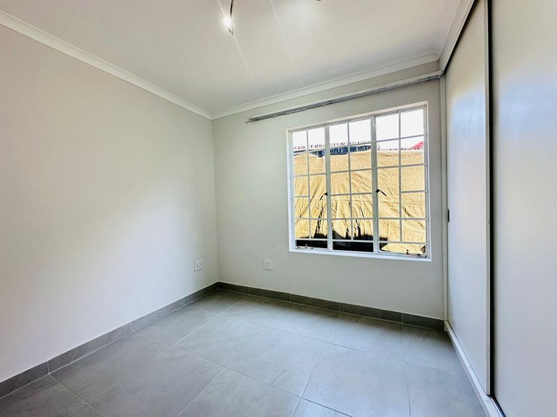 A clean, empty bedroom featuring neutral tiled flooring, a built-in wardrobe, and a large grid-pattern window providing natural light.