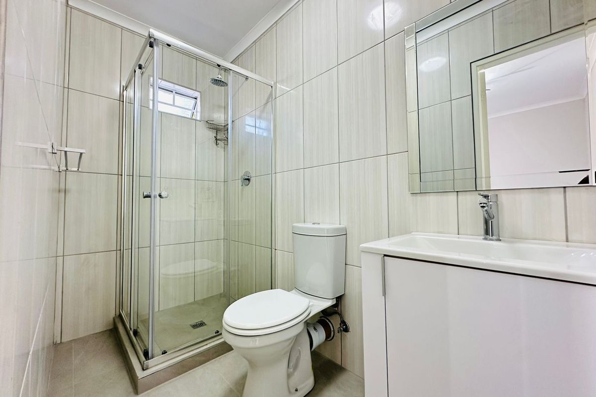 A clean, modern bathroom featuring a glass-enclosed shower, a white vanity with basin, and neutral floor-to-ceiling wall tiles.