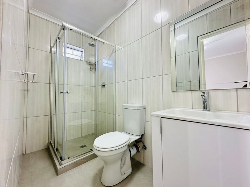 A clean, modern bathroom featuring a glass-enclosed shower, a white vanity with basin, and neutral floor-to-ceiling wall tiles.