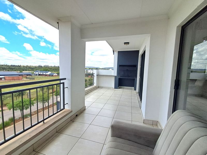 A spacious covered balcony featuring tiled flooring, a built-in braai, and metal balustrades, offering pleasant views of the surrounding estate.