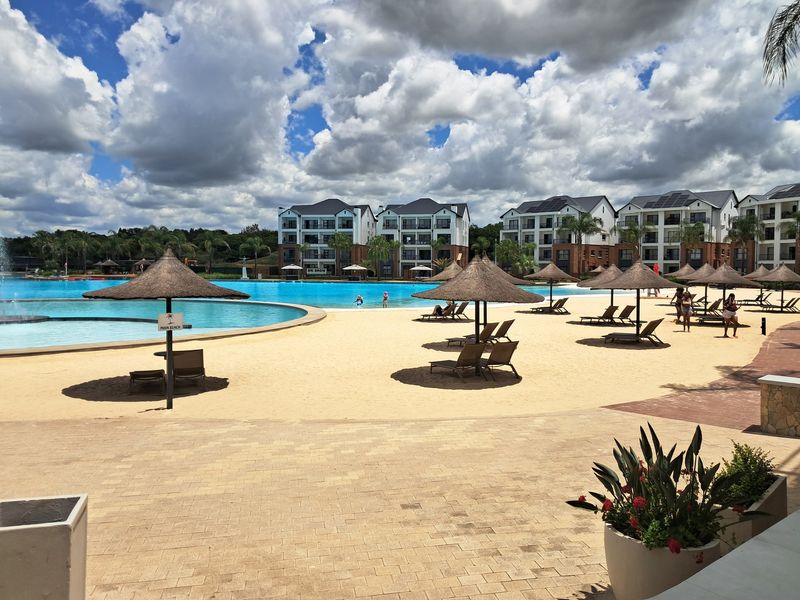 A large communal swimming pool area featuring a sandy beach, thatched umbrellas, and paved terrace, set within a modern apartment complex.