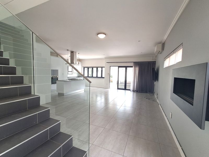 Spacious open-plan living area featuring modern tiled floors, a sleek kitchen island, built-in fireplace, and a contemporary glass-railed staircase leading to the upper level.