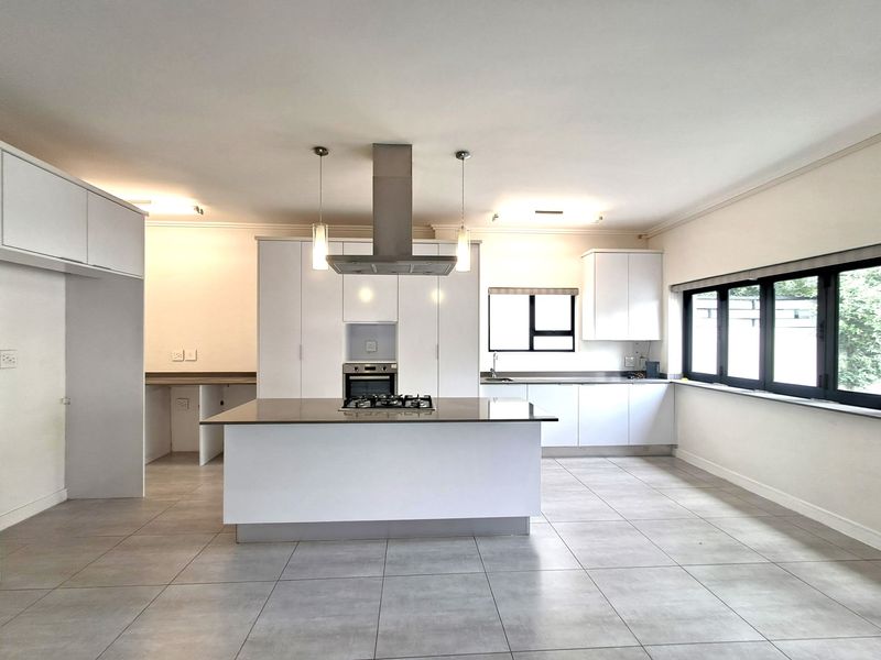 Modern, spacious kitchen featuring a central island with gas hob, built-in oven, sleek white cabinetry, and large windows providing ample natural light.