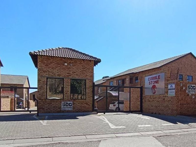 Front entrance view of the Cobble Stone apartment complex featuring a brick security gatehouse, paved driveway, and perimeter electric fencing.