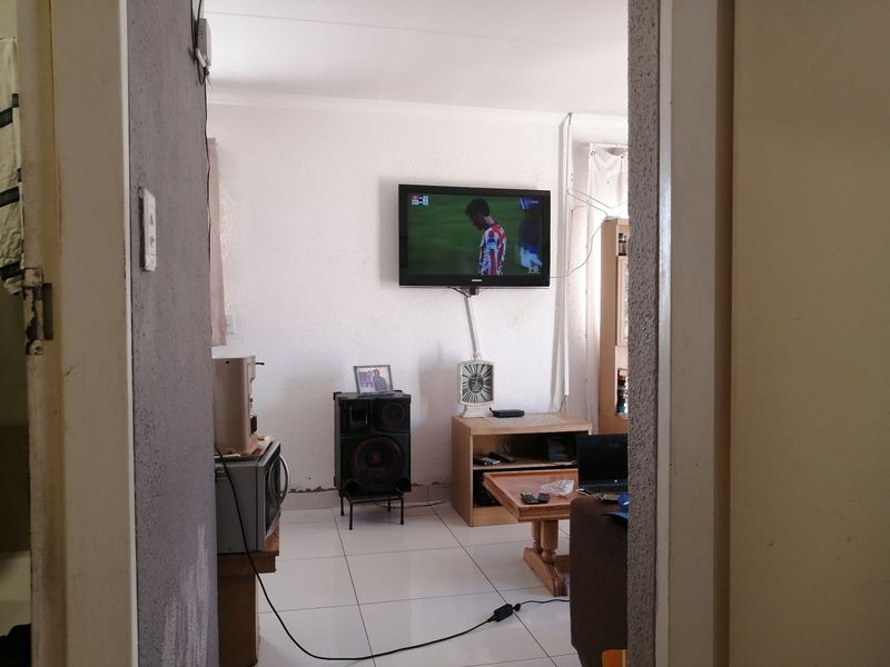 A living room area featuring tiled flooring and a wall-mounted television, viewed through a doorway.