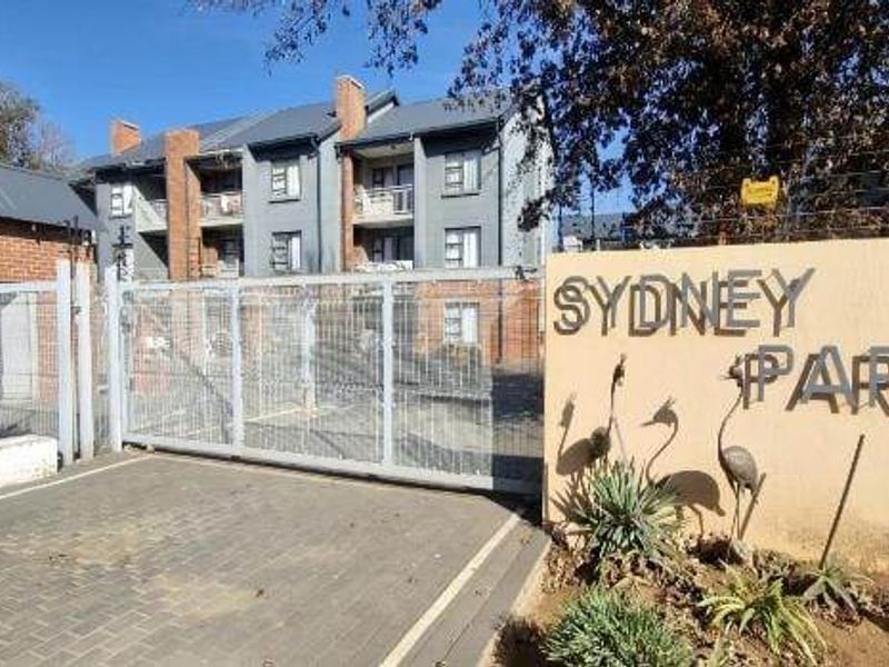 Front entrance view of Sydney Park apartment complex featuring a secure gated access point, paved driveway, and brick boundary wall.