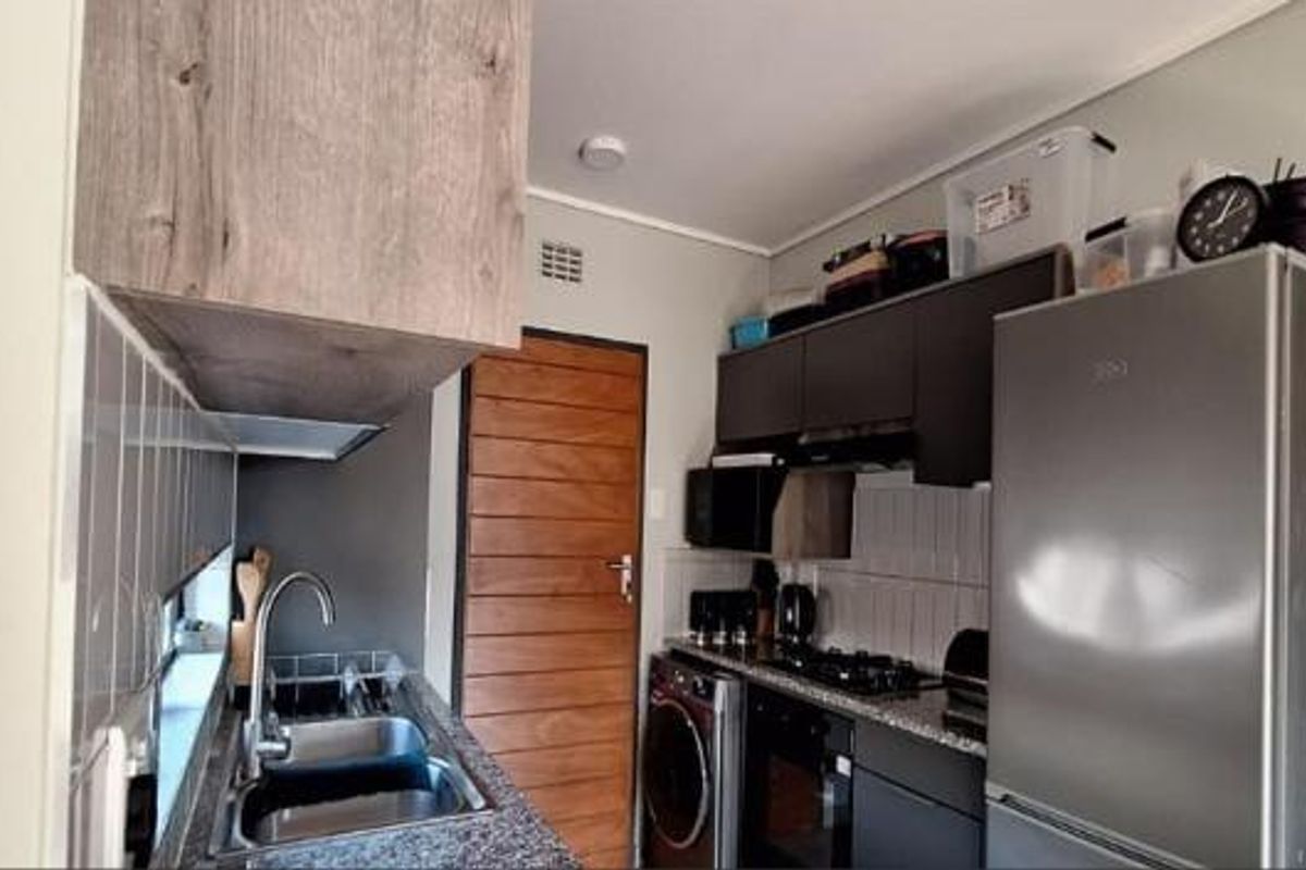 Compact modern kitchen featuring dark cabinetry, granite countertops, integrated oven, hob, extractor fan, and a stainless steel sink with tiled backsplash.