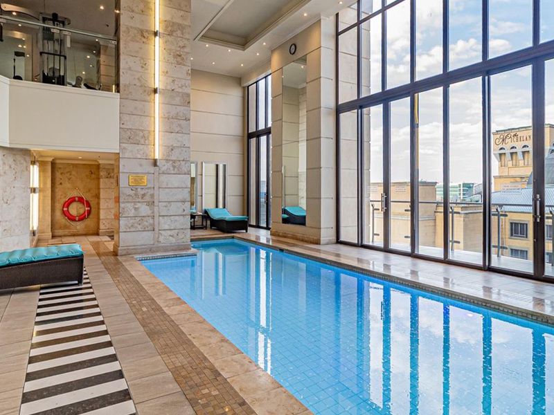 A luxurious indoor swimming pool area featuring high ceilings, stone-clad pillars, and expansive floor-to-ceiling windows offering an elevated urban view.