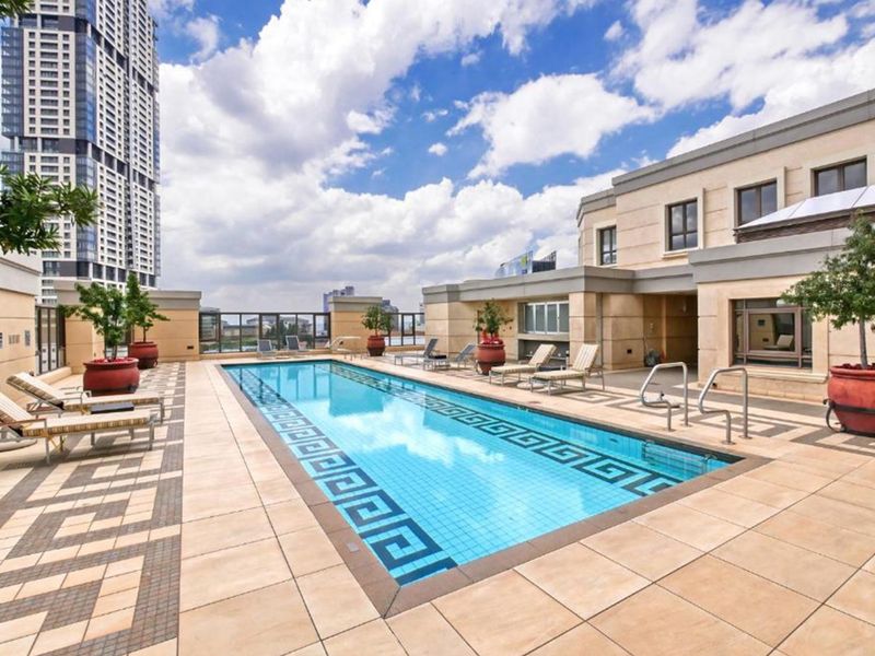 A well-maintained rooftop swimming pool area featuring tiled decking, glass balustrades, and a clear view of the surrounding urban skyline.