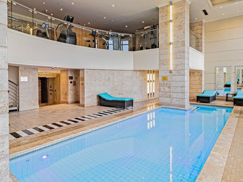 Luxury indoor swimming pool area featuring stone wall finishes, a mezzanine level with gym equipment, and modern recessed lighting.