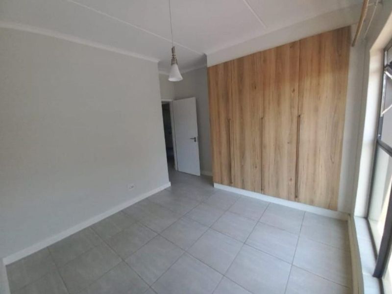 A bright, empty bedroom featuring modern wood-finish built-in cupboards, neutral tiled flooring, and a clean, minimalist aesthetic.