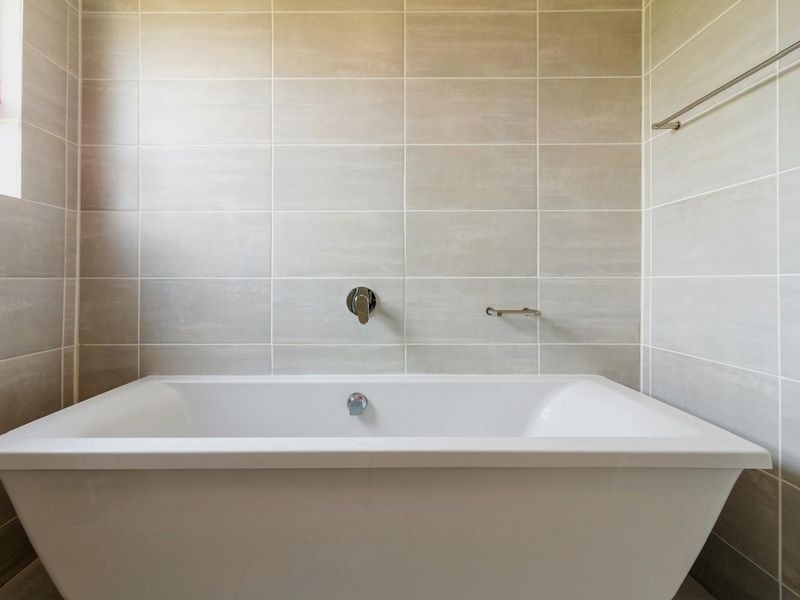 A clean, modern bathroom featuring a white bathtub with wall-mounted mixer tap and neutral ceramic wall tiling.