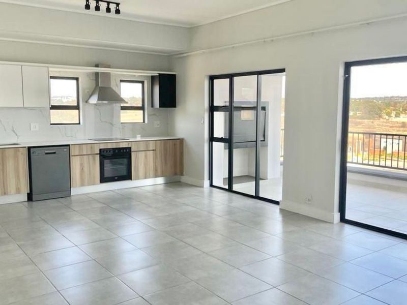 Modern open-plan kitchen and living area featuring sleek cabinetry, stone countertops, integrated appliances, and large sliding doors leading to a balcony with a built-in braai.