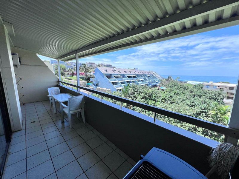 A spacious covered balcony featuring tiled flooring and a metal balustrade, offering expansive views of the surrounding coastal landscape and ocean.