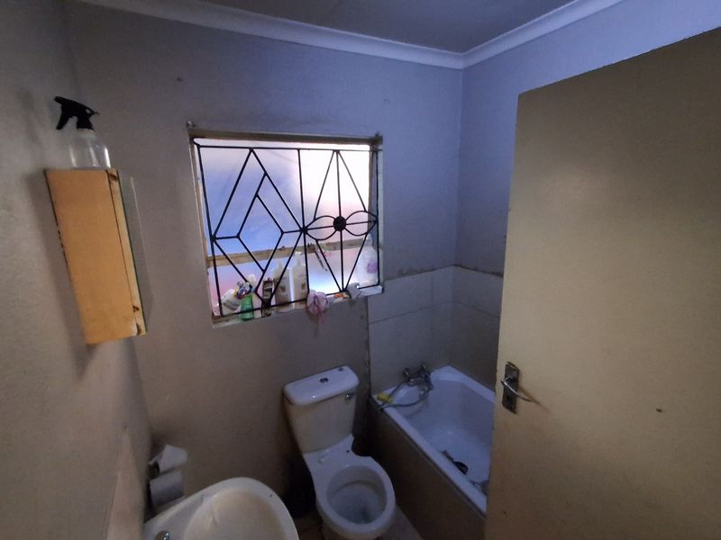 A compact bathroom featuring a bathtub, toilet, and basin, with security bars on the window and visible wear on the wall finishes.