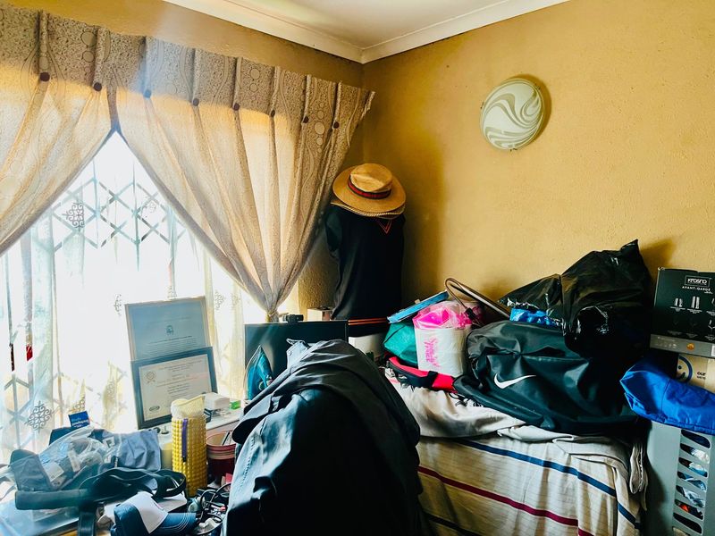 A bedroom featuring a window with security gate and ceiling mounted lighting, currently cluttered with personal belongings.