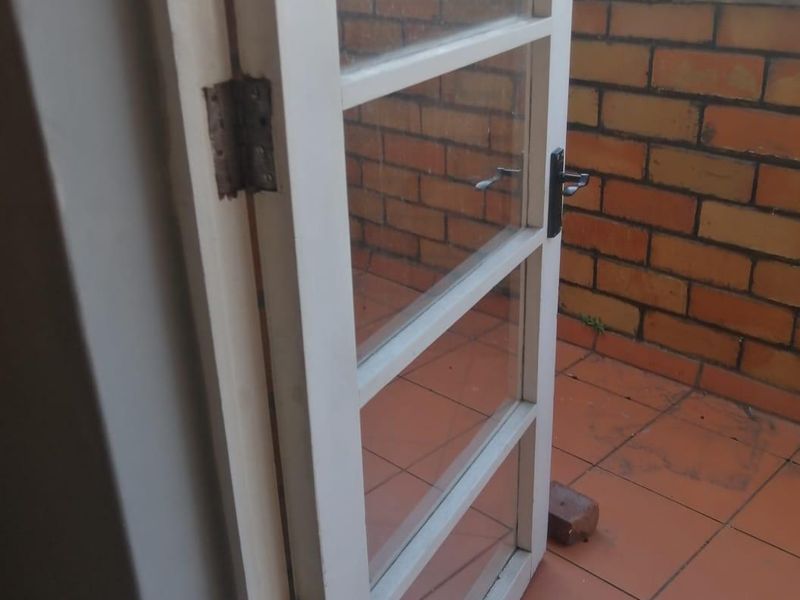 A balcony featuring terracotta floor tiles, a brick parapet wall, and a glass-paneled door with visible wear on the hinges.