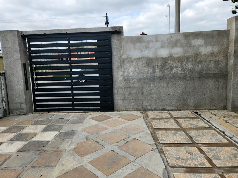 Front entrance area featuring a modern black sliding gate, concrete boundary wall, and a multi-patterned tiled driveway.