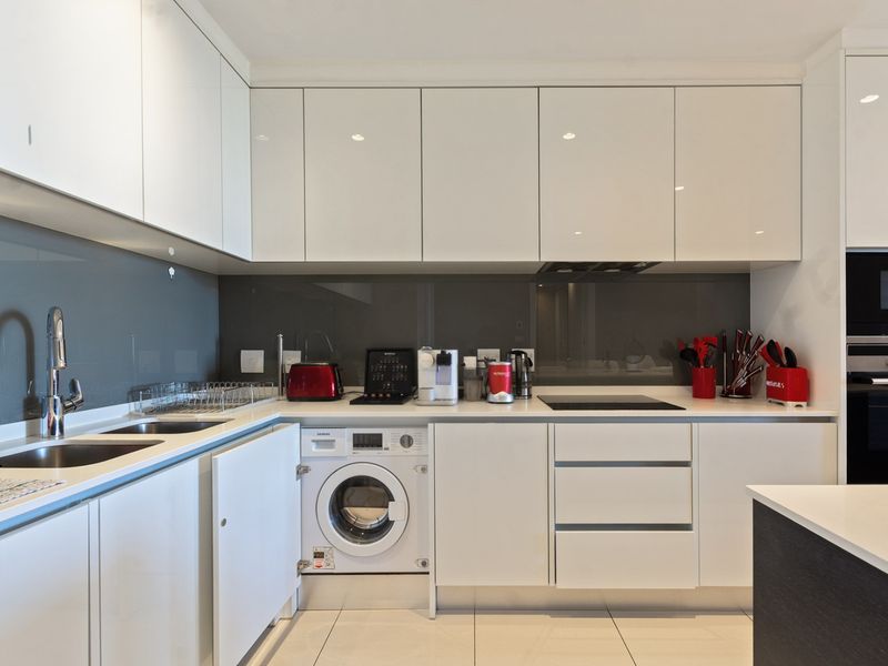 Modern kitchen featuring high-gloss white cabinetry, integrated appliances including a washing machine, oven, and microwave, complemented by stone countertops and a sleek glass splashback.