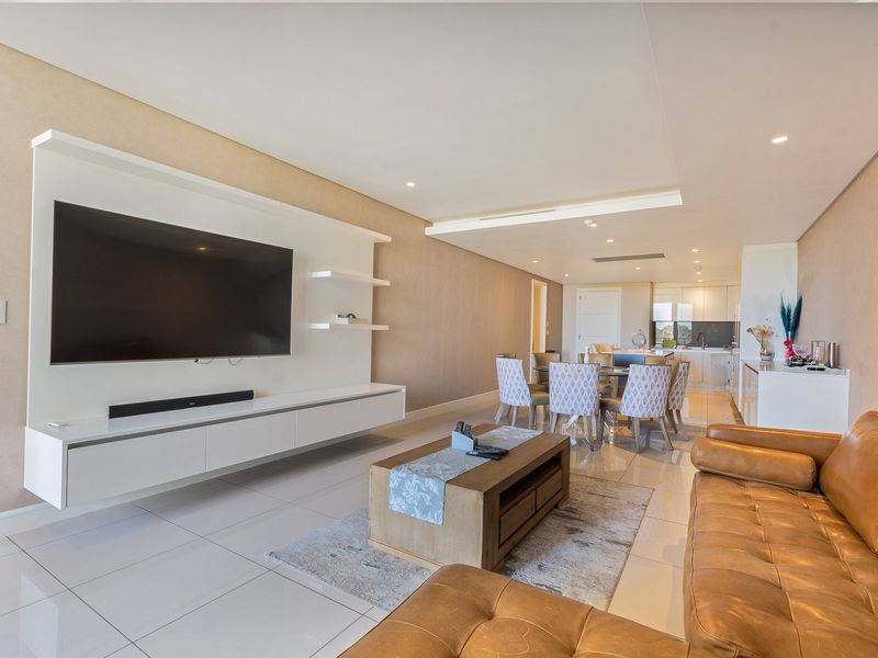 Spacious open-plan living and dining area featuring modern tiled floors, recessed lighting, and a sleek built-in media wall, flowing into a contemporary kitchen.