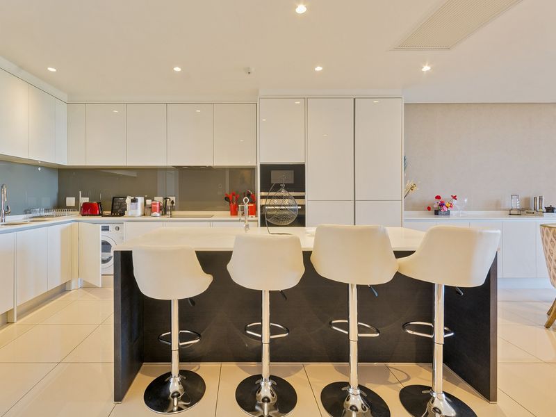 Modern open-plan kitchen featuring high-gloss white cabinetry, a central island with breakfast bar seating, integrated appliances, and sleek tiled flooring.