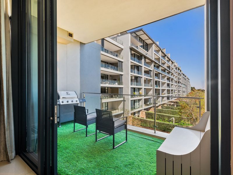 A modern covered balcony featuring a glass balustrade, stainless steel handrail, and recessed ceiling lighting, overlooking the apartment complex grounds.