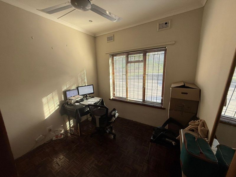 A functional office space featuring parquet flooring, a ceiling fan, and large windows with venetian blinds, offering a versatile area for work or study.