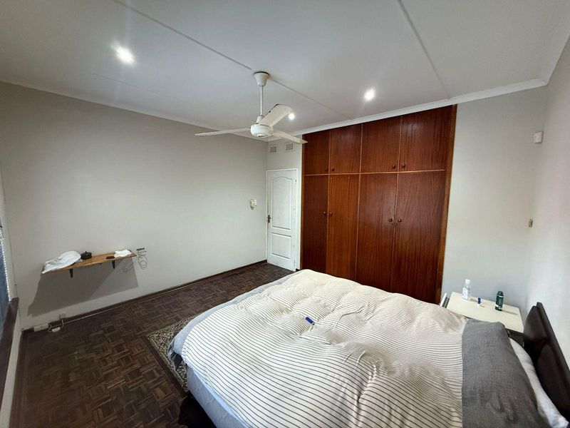 A bedroom featuring classic parquet flooring, built-in wooden cupboards, and a ceiling fan, offering a functional space with potential for modern updates.
