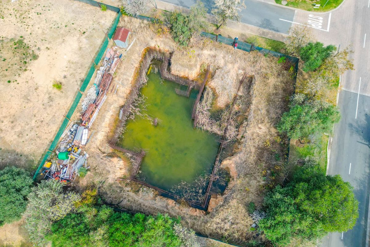Aerial view of a vacant residential plot featuring an excavated foundation pit with stagnant water and exposed rebar reinforcement.