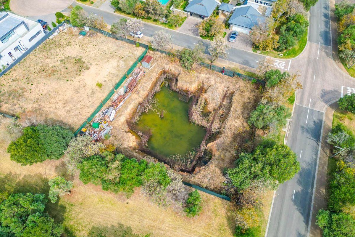 Aerial view of a vacant residential plot featuring an excavated foundation pit with stagnant water and exposed steel reinforcement bars.