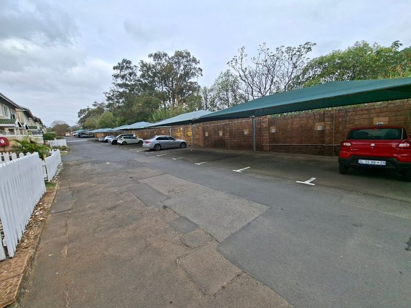 Outdoor parking area featuring covered carports with shade netting, set against a boundary wall and paved driveway within the townhouse complex.