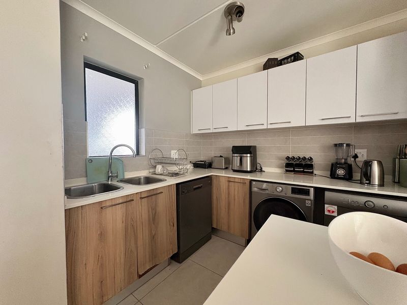 A functional kitchen featuring wood-look lower cabinetry, white upper cabinets, stone countertops, a double sink, and integrated laundry and dishwasher appliances.