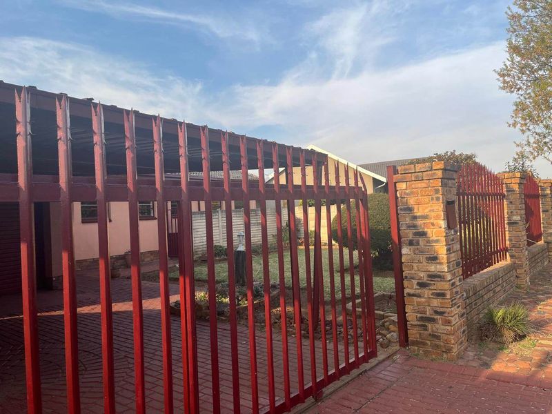 Front view of the property featuring a paved driveway, brick boundary wall with pillars, and a red steel security gate.