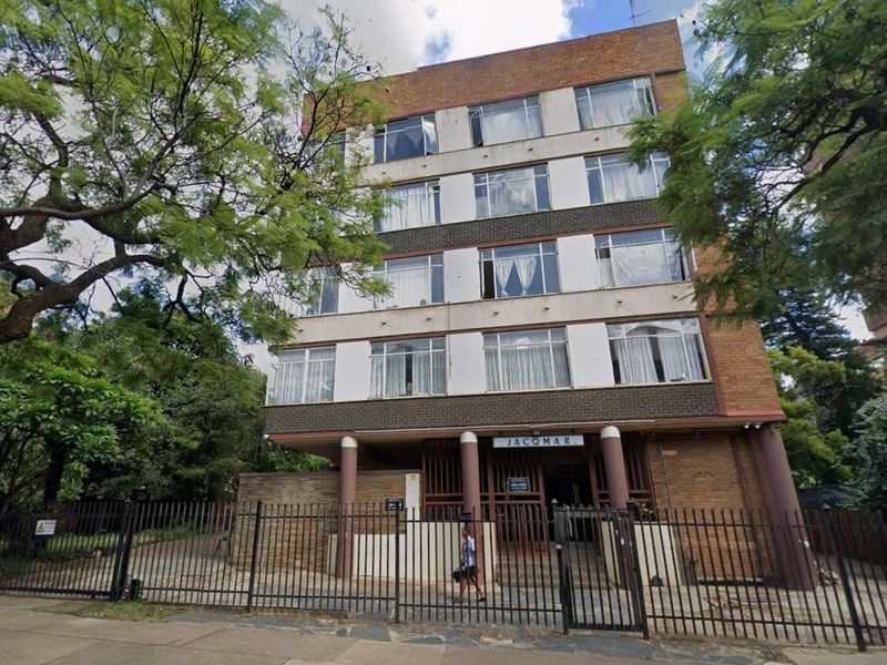 Front elevation of the Jacqmar apartment building, featuring a multi-story brick facade, metal security fencing, and established surrounding trees.