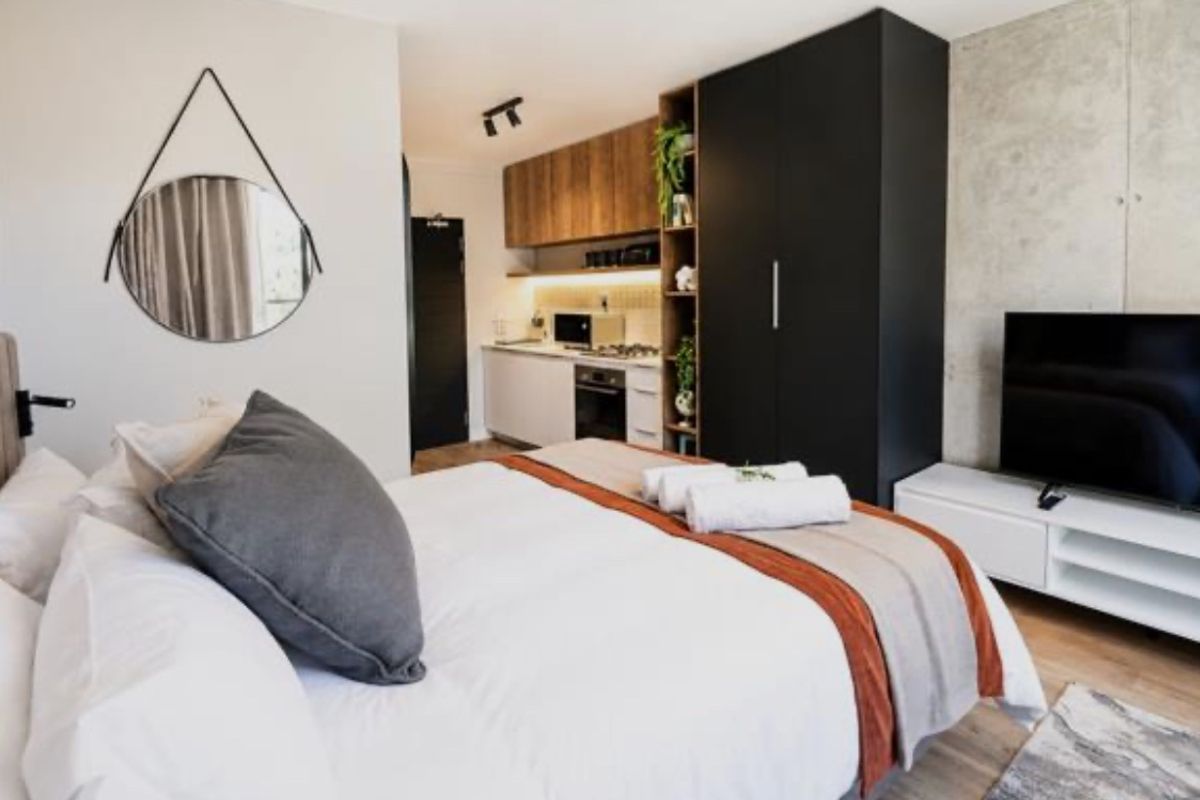 Modern studio bedroom featuring a compact integrated kitchen with built-in cabinetry, oven, and hob, complemented by a stylish concrete feature wall.