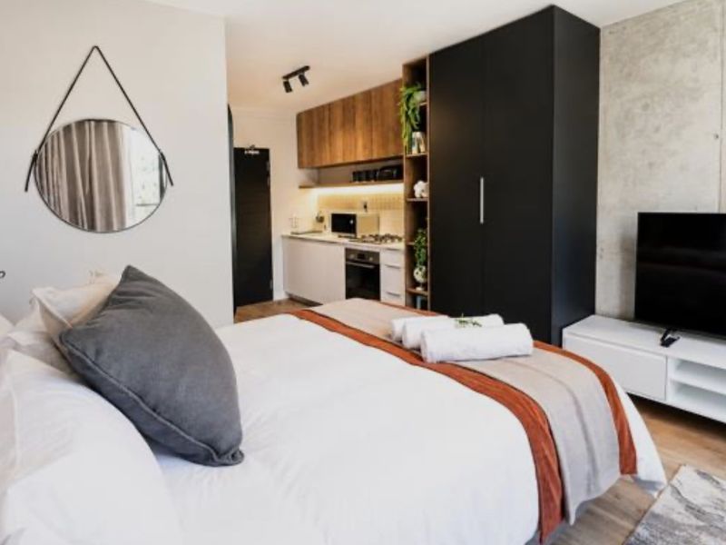Modern studio bedroom featuring a compact integrated kitchen with built-in cabinetry, oven, and hob, complemented by a stylish concrete feature wall.