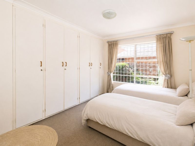 A bedroom featuring extensive built-in cupboards, neutral carpeting, and a window fitted with security bars overlooking the garden.