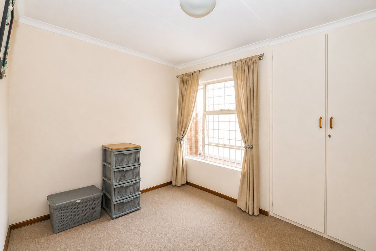A clean, neutral bedroom featuring built-in cupboards, carpeted flooring, and a window fitted with security bars.