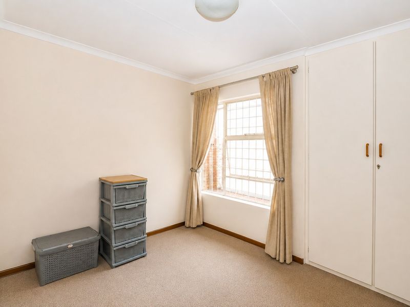 A clean, neutral bedroom featuring built-in cupboards, carpeted flooring, and a window fitted with security bars.