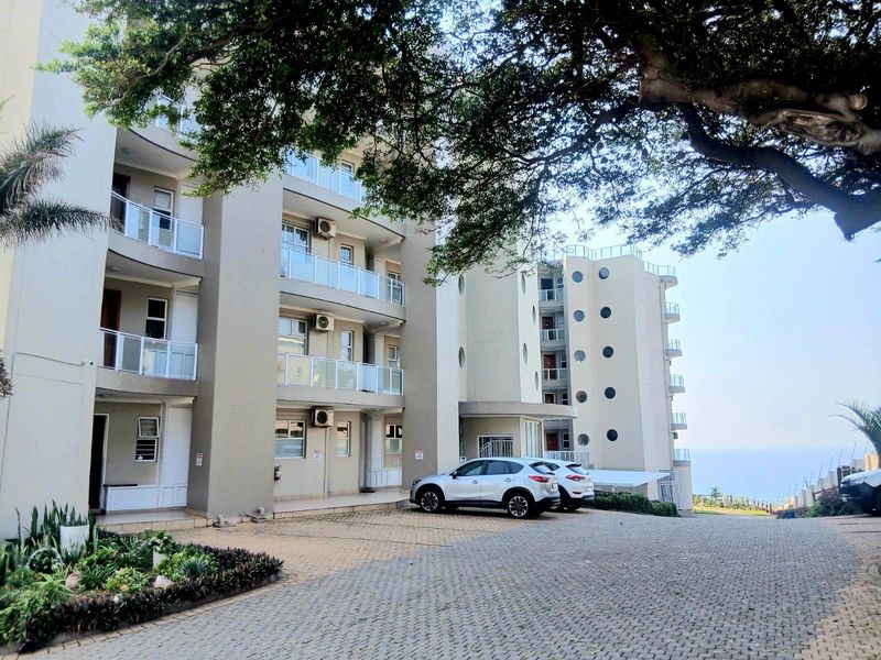 Exterior view of a multi-story apartment complex featuring paved parking, balconies with glass balustrades, and distinctive porthole windows overlooking the coast.
